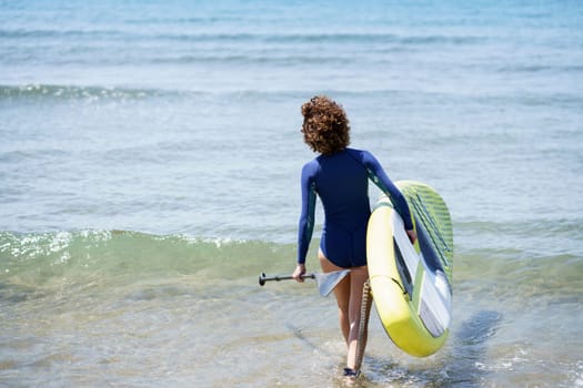 Back view of unrecognizable female surfer in wetsuit walking on shallow beach of waving sea while carrying SUP board and paddle in daylight