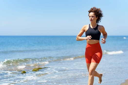 Concentrated barefoot young female with curly hair, wearing sportswear and looking away while jogging in daylight on sandy beach near wavy seawater against blurred blue sky