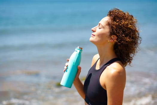 Side view of positive female athlete in sportswear with closed eyes standing with bottle of fresh water and enjoying sea breeze