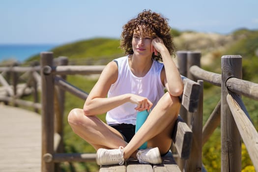 Positive young female in sportswear sitting on wooden fence and looking away while relaxing with crossed legs against blurred background water bottle
