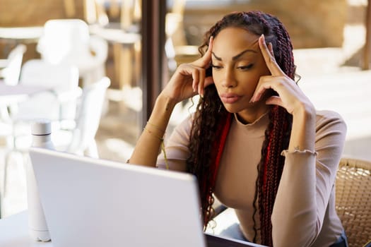 A woman sporting beautifully braided hair sits deep in thought, intensely focused on her laptop while immersed in the vibrant ambiance of a stylish cafe, blending work with casual elegance