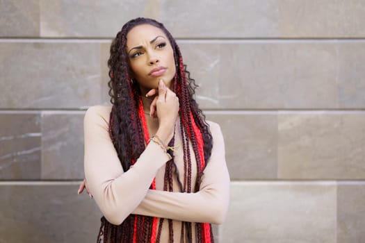 A woman stands thoughtfully against an intriguing textured wall, showcasing her distinctive braided hairstyle adorned with vibrant red accents, embodying confidence, creativity, and personal style