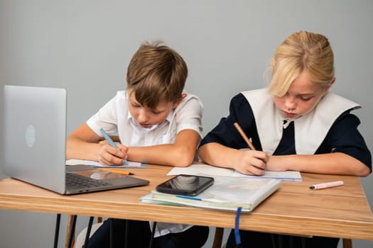 Two children, a boy and a girl, are seated at a table doing schoolwork. A laptop, pencil, and notebook are on the table.