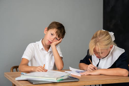 A bored boy rests his head on his hand while a girl diligently writes in a notebook at a wooden desk.