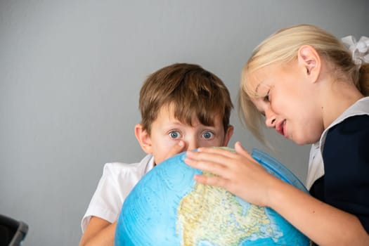 Two children, a boy and a girl, wearing school uniforms, look intently at a globe. The boy is touching the globe with his finger, and the girl is looking at it with a serious expression.