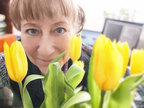 A woman with bright blue eyes smiles warmly while holding a bouquet of fresh yellow tulips in a lively indoor environment.