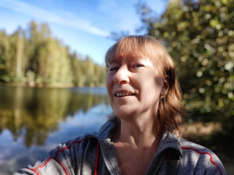A middle-aged woman takes a cheerful selfie by a tranquil lake surrounded by lush trees, capturing a moment of joy in nature.
