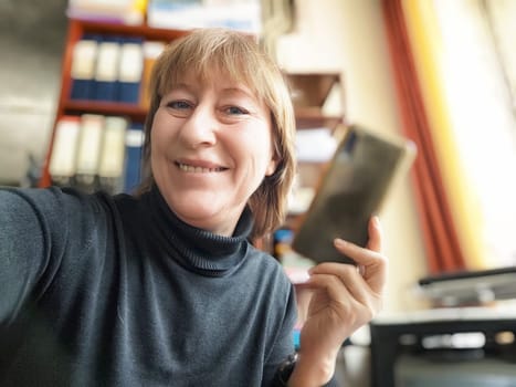 A mature woman smiles brightly as she holds her phone, capturing a joyful selfie in her inviting blogging space filled with travel inspirations.