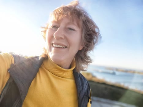 A mature woman enjoys a bright day outdoors, smiling happily for a selfie while exploring a scenic destination near the water.