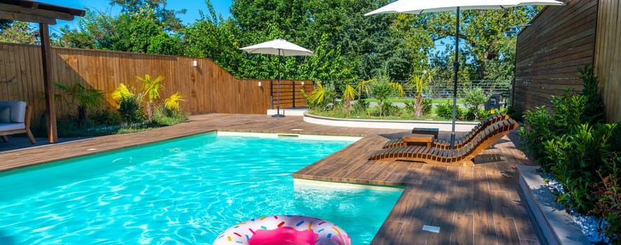Modern backyard swimming pool with turquoise water, wooden deck, lounge chairs, umbrellas, and pink inflatable ring