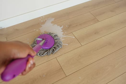 A person mops a light wooden floor with a purple mop in a sunny space.