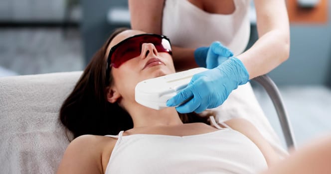 Beauty spa providing laser epilation treatment for young woman's cheek. Professional beautician.