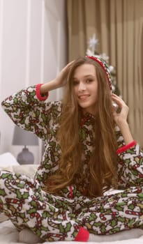 Beautiful young woman in pajamas sits on the bed against the background of a Christmas tree. She smiles and makes wishes. Christmas and New Year concept. Looking at the camera.