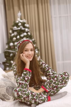 Beautiful young woman in pajamas sits on the bed against the background of a Christmas tree. She smiles and makes wishes. Christmas and New Year concept. Looking at the camera