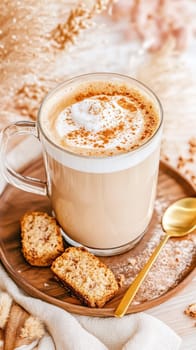 A creamy latte garnished with cinnamon sits in a clear glass mug alongside two oat biscuits on a wooden tray. The cozy setup suggests warmth and comfort, perfect for a relaxed afternoon.