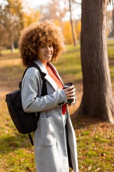 A joyful woman with curly hair strolls through a sunny park adorned with colorful autumn leaves, holding a warm cup of coffee while wearing a stylish outfit.