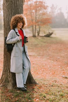 A young woman with curly hair leans against a sturdy tree, holding a warm drink. Dressed in a cozy grey coat, she enjoys the tranquil atmosphere of an autumn day.