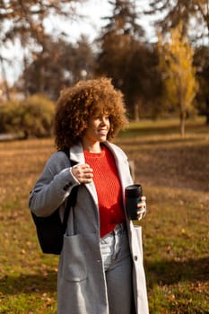 A joyful woman with curly hair strolls through a sunny park adorned with colorful autumn leaves, holding a warm cup of coffee while wearing a stylish outfit.