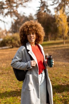 A joyful woman with curly hair strolls through a sunny park adorned with colorful autumn leaves, holding a warm cup of coffee while wearing a stylish outfit.