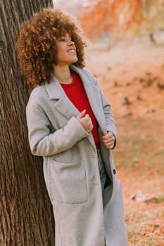 A young woman with curly hair leans against a tree, wearing a warm sweater, smiling joyfully amidst the golden autumn foliage and fallen leaves around her.