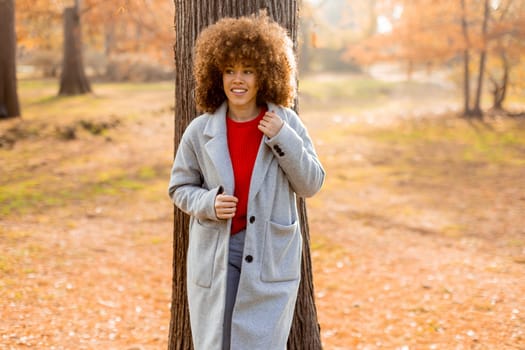 A young woman with curly hair leans against a tree, wearing a warm sweater, smiling joyfully amidst the golden autumn foliage and fallen leaves around her.