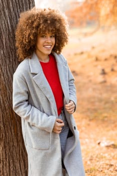A young woman with curly hair leans against a tree, wearing a warm sweater, smiling joyfully amidst the golden autumn foliage and fallen leaves around her.