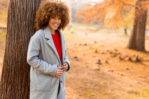 A young woman with curly hair leans against a tree, wearing a warm sweater, smiling joyfully amidst the golden autumn foliage and fallen leaves around her.