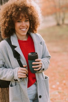 A young woman with curly hair leans against a sturdy tree, holding a warm drink. Dressed in a cozy grey coat, she enjoys the tranquil atmosphere of an autumn day.