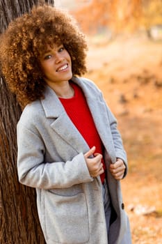 A young woman with curly hair leans against a tree, wearing a warm sweater, smiling joyfully amidst the golden autumn foliage and fallen leaves around her.