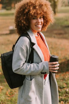 A cheerful woman strolls through a vibrant autumn park wearing a cozy sweater and stylish coat while holding a coffee cup, surrounded by colorful fallen leaves.
