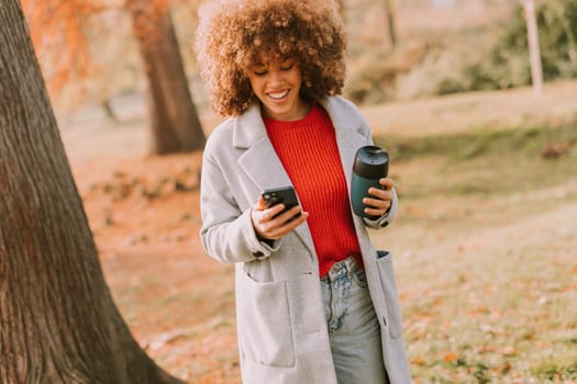 A young woman with curly hair wears a cozy sweater and a stylish coat while walking through a vibrant autumn park, happily engaged with her phone and coffee.