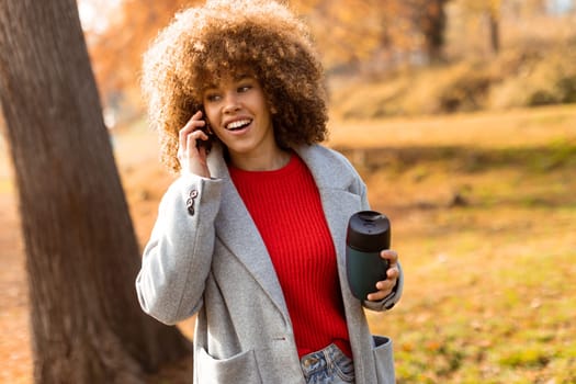Bright autumn foliage surrounds a young woman, who is smiling and conversing on her phone, with a warm beverage in hand. She embodies a joyful moment in nature.
