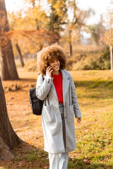 A young woman with curly hair smiles brightly while talking on her phone, dressed warmly in a stylish coat amidst a picturesque autumn landscape filled with soft light.