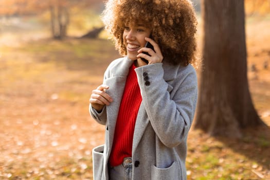 With a beaming smile, a young woman with curly hair engages in a warm conversation on her phone while enjoying the colorful autumn landscape in the park, radiating happiness.