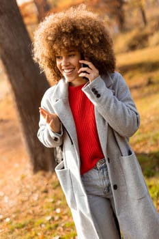 With a beaming smile, a young woman with curly hair engages in a warm conversation on her phone while enjoying the colorful autumn landscape in the park, radiating happiness.