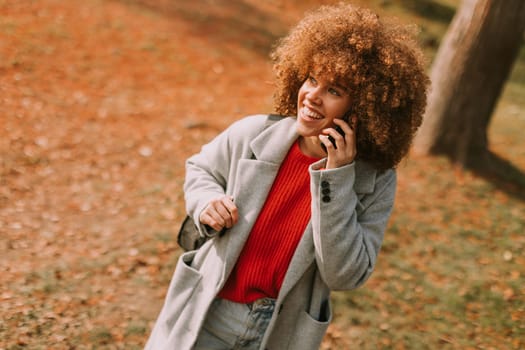 A young woman with curly hair laughs while talking on her phone, dressed in a stylish gray coat and vibrant red sweater, surrounded by the warm hues of autumn foliage.