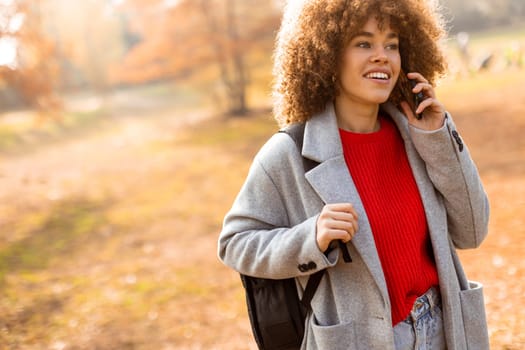 A young woman with curly hair laughs while talking on her phone, dressed in a stylish gray coat and vibrant red sweater, surrounded by the warm hues of autumn foliage.