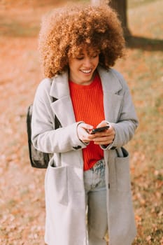 A joyful woman with curly locks is engaging with her smartphone, surrounded by the warm colors of autumn leaves in a lively outdoor park setting.