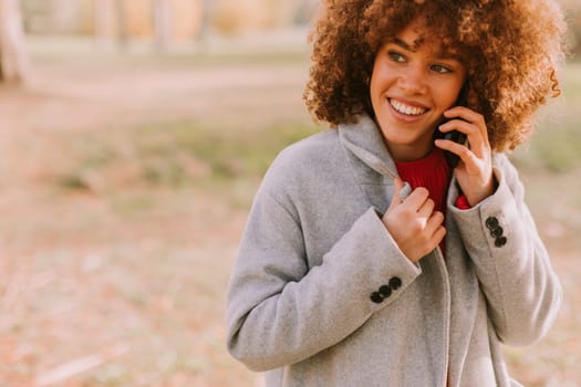A young woman with curly hair smiles brightly while talking on her phone, dressed warmly in a stylish coat amidst a picturesque autumn landscape filled with soft light.