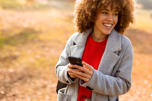 A cheerful individual with curly hair smiles brightly as she engages with her smartphone, wearing a cozy red sweater and a stylish gray coat amidst golden autumn foliage.