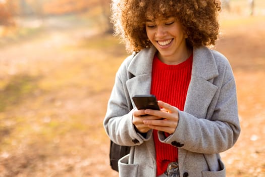 A cheerful individual with curly hair smiles brightly as she engages with her smartphone, wearing a cozy red sweater and a stylish gray coat amidst golden autumn foliage.