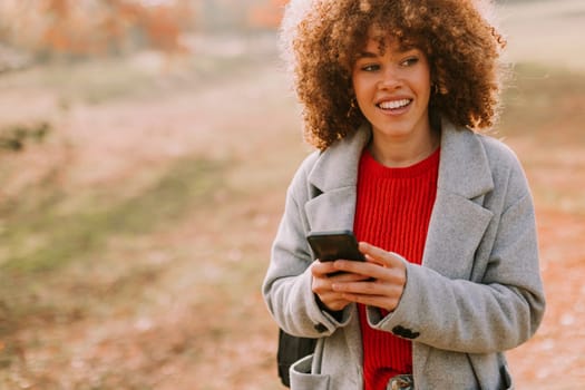 A cheerful individual with curly hair smiles brightly as she engages with her smartphone, wearing a cozy red sweater and a stylish gray coat amidst golden autumn foliage.