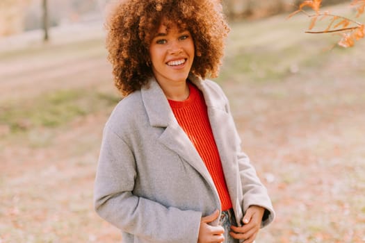 A joyful woman with curly hair stands in a lush park, dressed in a gray coat and bright red sweater. Golden autumn leaves create a vibrant backdrop as she smiles warmly.