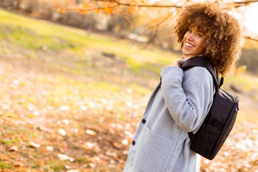 In a beautiful autumn park, a joyful young woman with curly hair showcases her bright smile while wearing a stylish grey coat and red sweater, carrying a backpack.