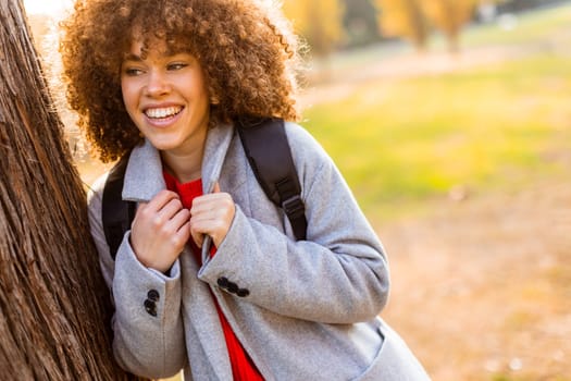 Smiling young woman with curly hair stands against a tree, wearing a cozy outfit. She embraces the beauty of autumn in a serene park, filled with colorful leaves and soft sunlight.