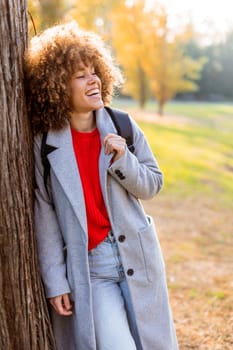 Smiling young woman with curly hair stands against a tree, wearing a cozy outfit. She embraces the beauty of autumn in a serene park, filled with colorful leaves and soft sunlight.