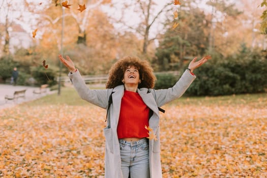 A cheerful individual with curly hair plays in a park during autumn, joyfully tossing vibrant leaves into the air among a golden blanket of fallen foliage.