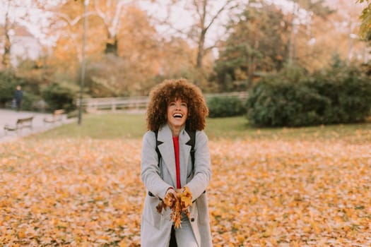 A cheerful individual with curly hair plays in a park during autumn, joyfully tossing vibrant leaves into the air among a golden blanket of fallen foliage.