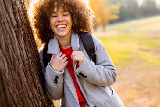 A carefree young woman laughs heartily while standing next to a tree in a serene park. The warm sunlight filters through the leaves, creating a joyful atmosphere.