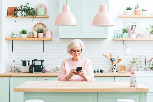 A joyful senior woman sits at a wooden kitchen island, engaged with her smartphone. The pastel-colored kitchen features stylish decor and plants, creating a cozy atmosphere.